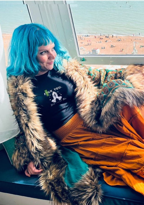 A person with bright blue hair reclines on a bench by a window, wearing a black top, a long orange skirt, and a brown fur-trimmed outer layer. The scene is set indoors near the window with an ocean view and beach visible outside.