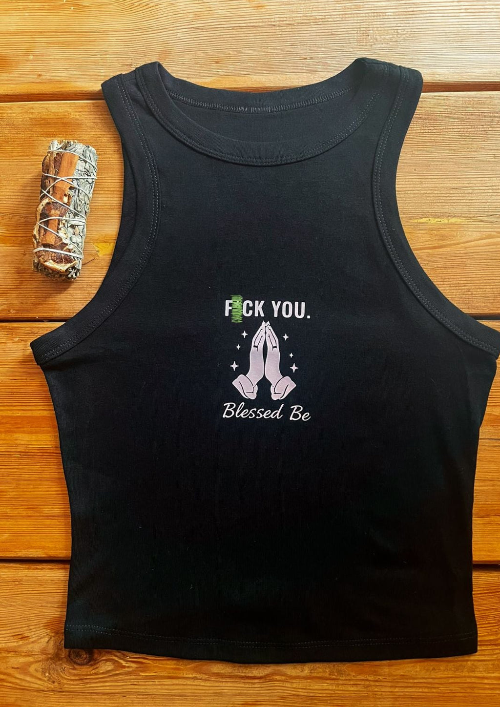 A black sleeveless tank top laid flat on a wooden surface, featuring a graphic print on the front with the text 'F YOU' in green and white, a pink praying girl illustration, and 'Blessed Be' in white script.