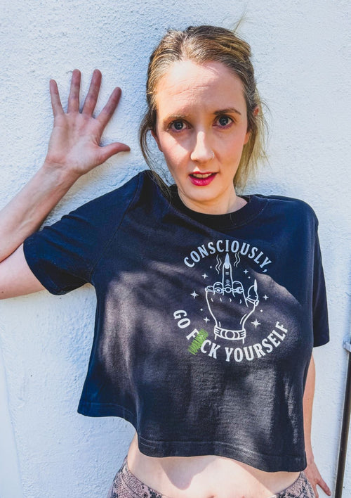A woman posing against a white textured wall wearing a black graphic crop top t-shirt with the text 'CONSCIOUSLY GO F YOURSELF' and a hand graphic.