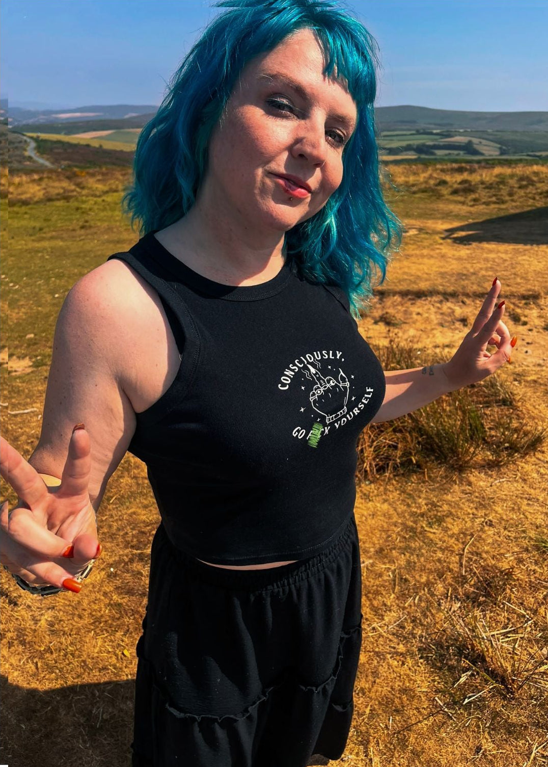 A woman with vibrant blue hair stands outdoors in a grassy field, wearing a black sleeveless crop top with a white graphic print that reads 'CONSCIOUSLY. GO F YOURSELF.