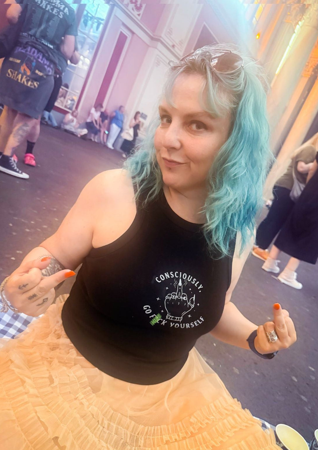 A woman with bright blue-green hair wearing a black tank top with a white graphic logo that says 'Consciously, go F Yourself' and a beige ruffled skirt, photographed in a crowded outdoor street setting. 