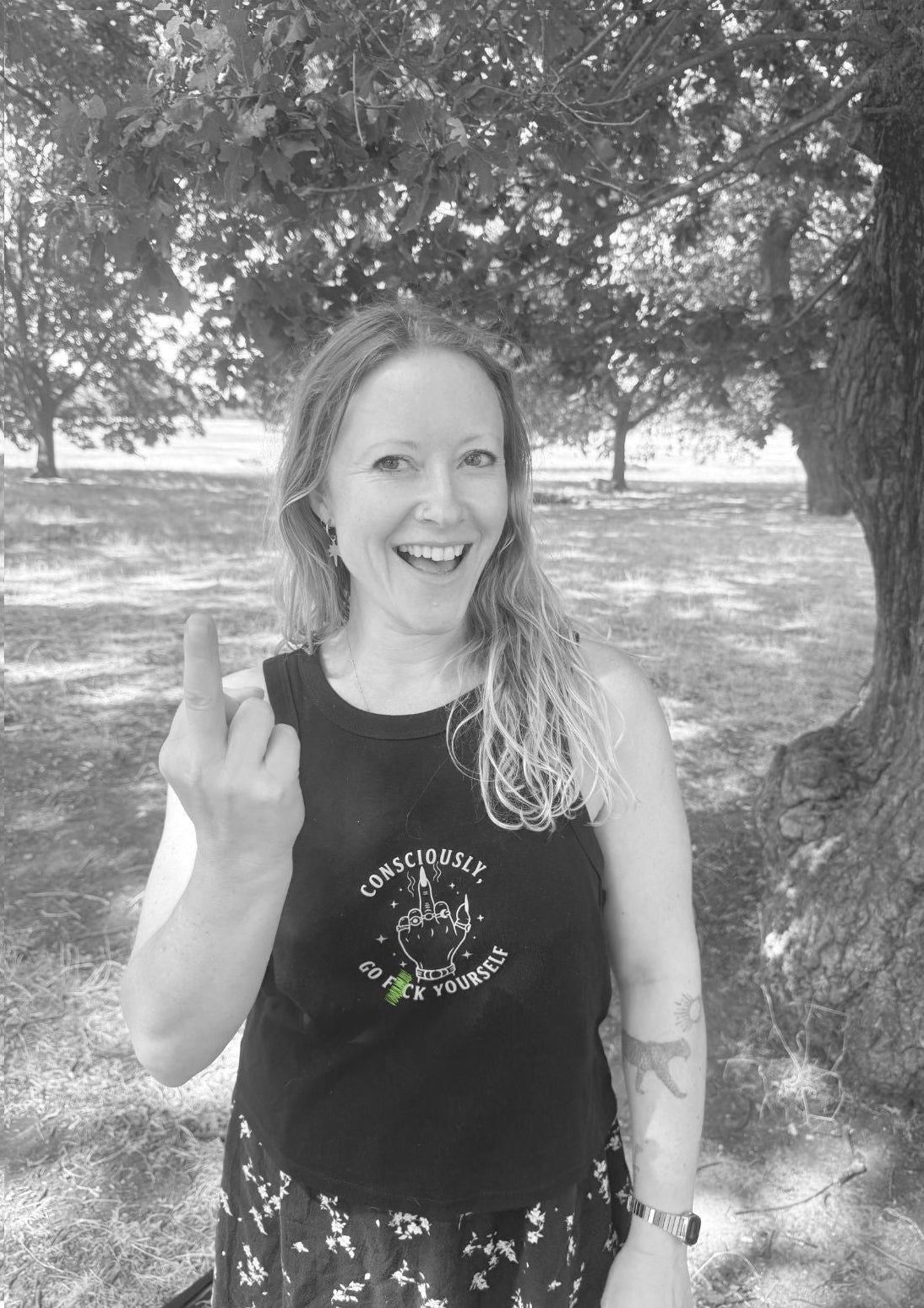 A woman outdoors under a tree wearing a black sleeveless tank top with a circular graphic and text on the front saying 'Consciously Go F yourself', paired with a dark floral skirt. She is holding up her middle finger.