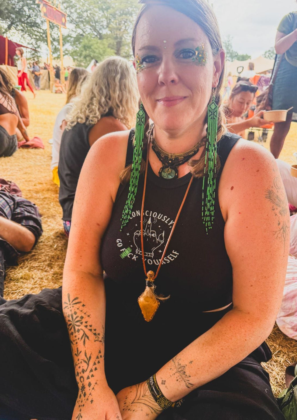 A woman with green braided hair sits outdoors at what looks like a festival or gathering. She is wearing a black sleeveless top with white text that reads 'Consciously, go F yourself', layered with multiple gold-toned necklaces, including one with a large pendant.