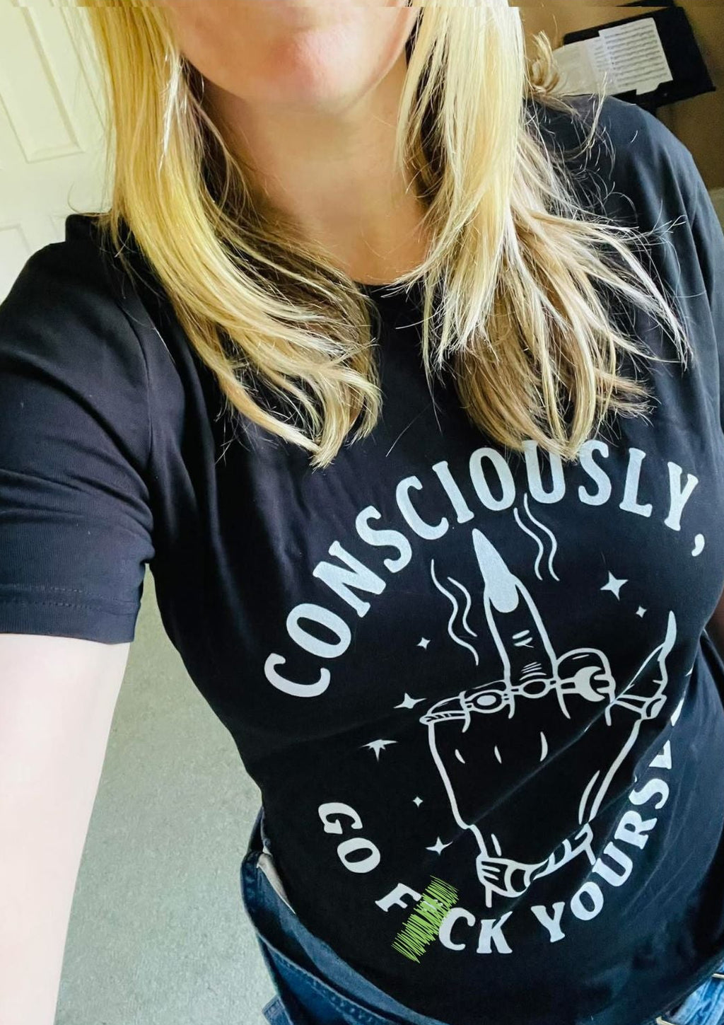 A woman wearing a black graphic t-shirt with a white hand-drawn style design and the text 'CONSCIOUSLY, GO F YOURSELF' in a casual indoor setting.