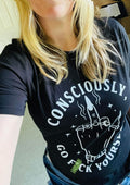A woman wearing a black graphic t-shirt with a white hand-drawn style design and the text 'CONSCIOUSLY, GO F YOURSELF' in a casual indoor setting.
