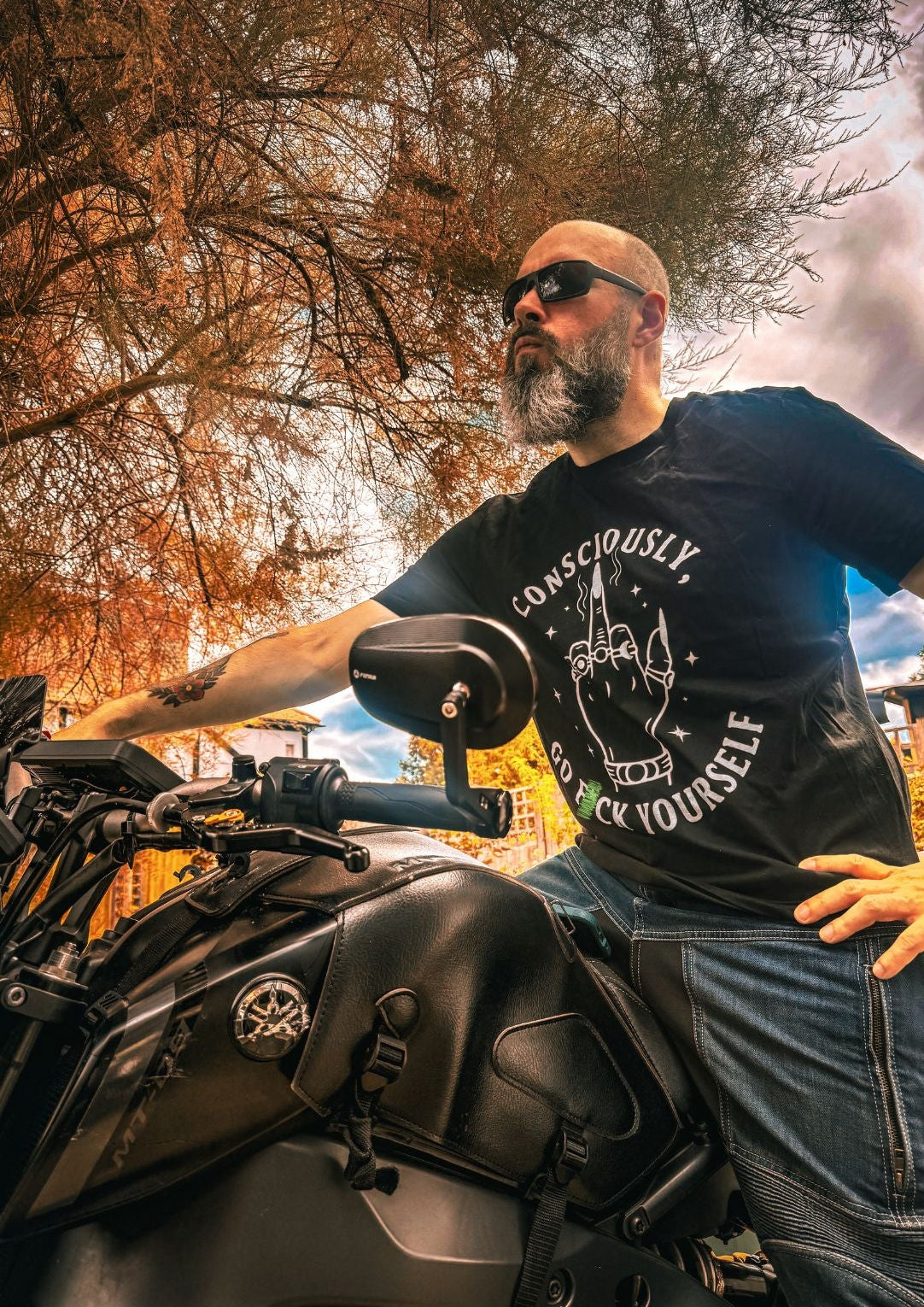 A bearded man wearing sunglasses and a black graphic t-shirt with a motorcycle, photographed from a low angle under autumn trees.
