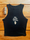 A black sleeveless tank top laid flat on a wooden surface, featuring a white printed text graphic that reads 'PSA The people pleaser has left the building' with a hand icon. A silver smudge stick is placed to the left of the tank top.