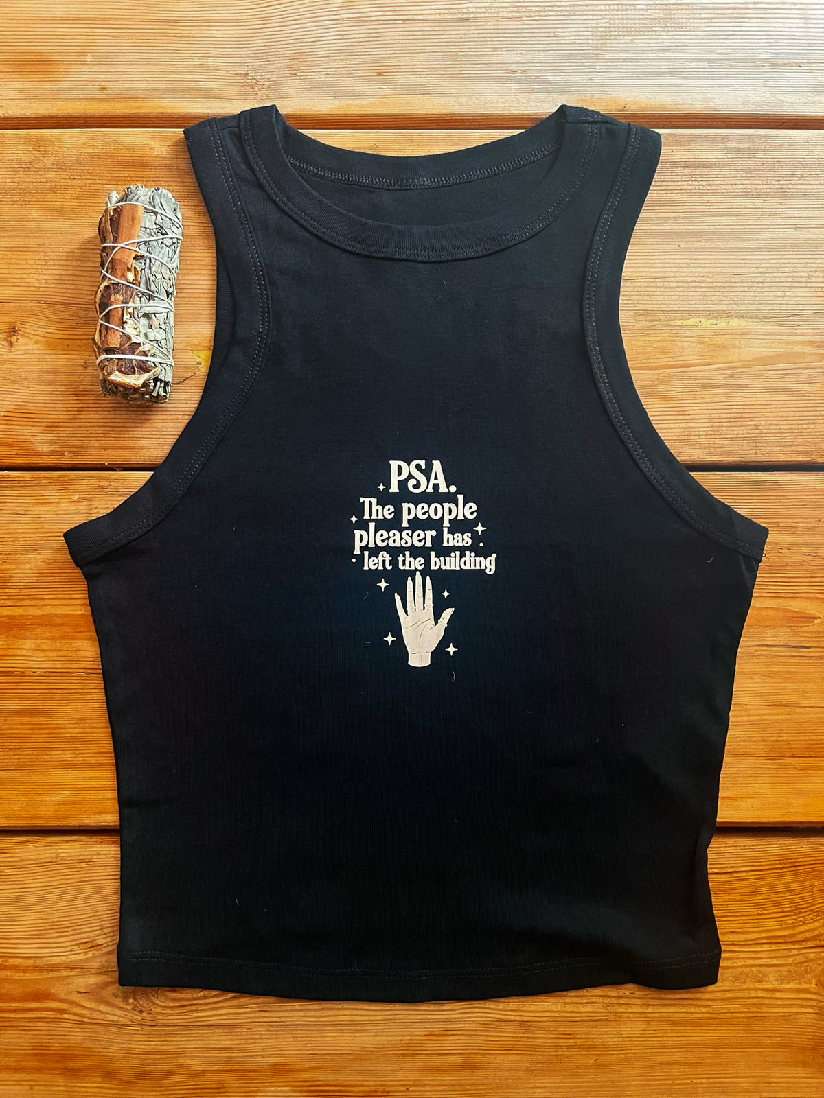A black sleeveless tank top laid flat on a wooden surface, featuring a white printed text graphic that reads 'PSA The people pleaser has left the building' with a hand icon. A silver smudge stick is placed to the left of the tank top.