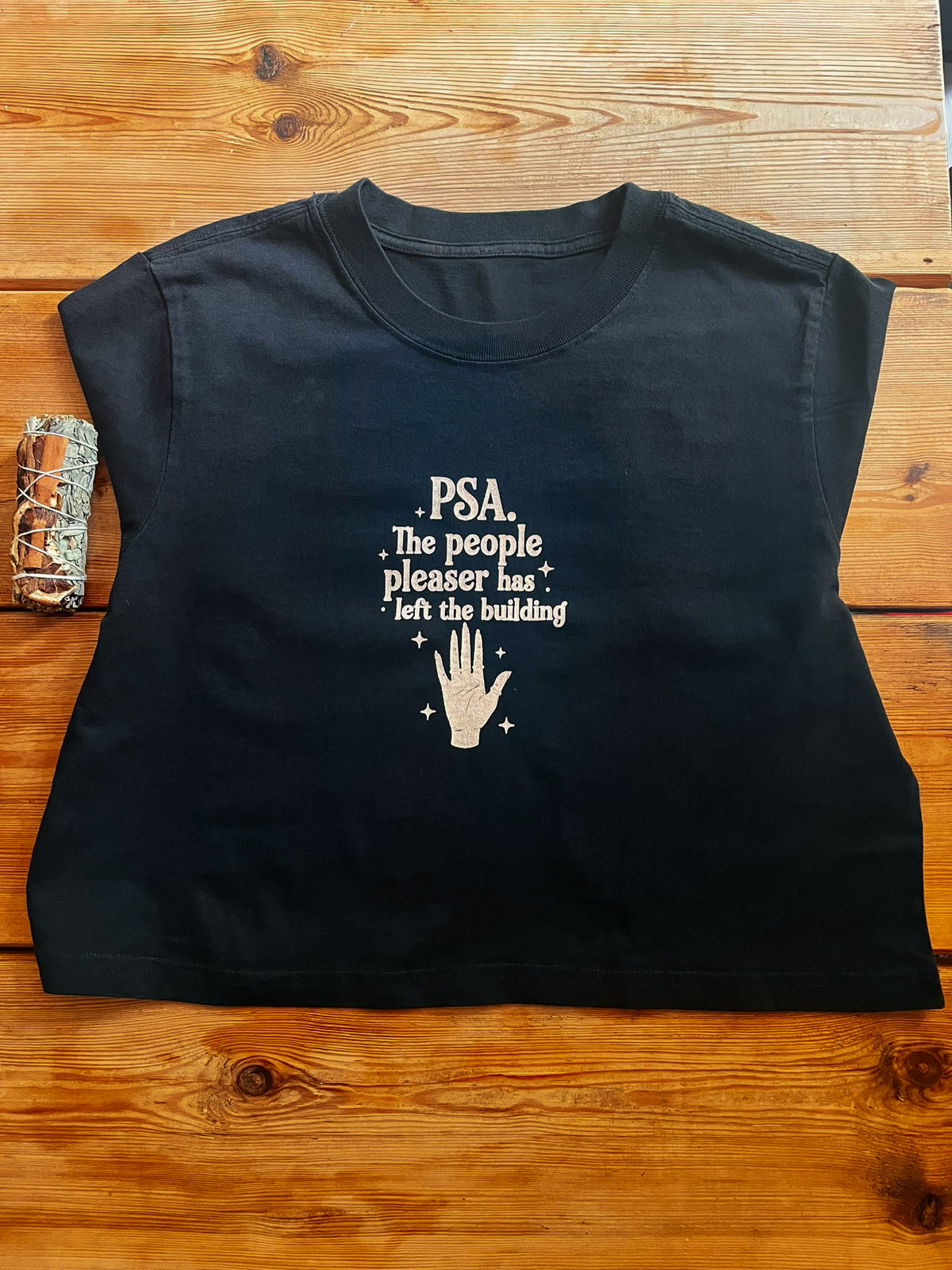 A black short-sleeve t-shirt with a white graphic print that reads 'PSA. The people pleaser has left the building' above a hand graphic, laid flat on a wooden surface.