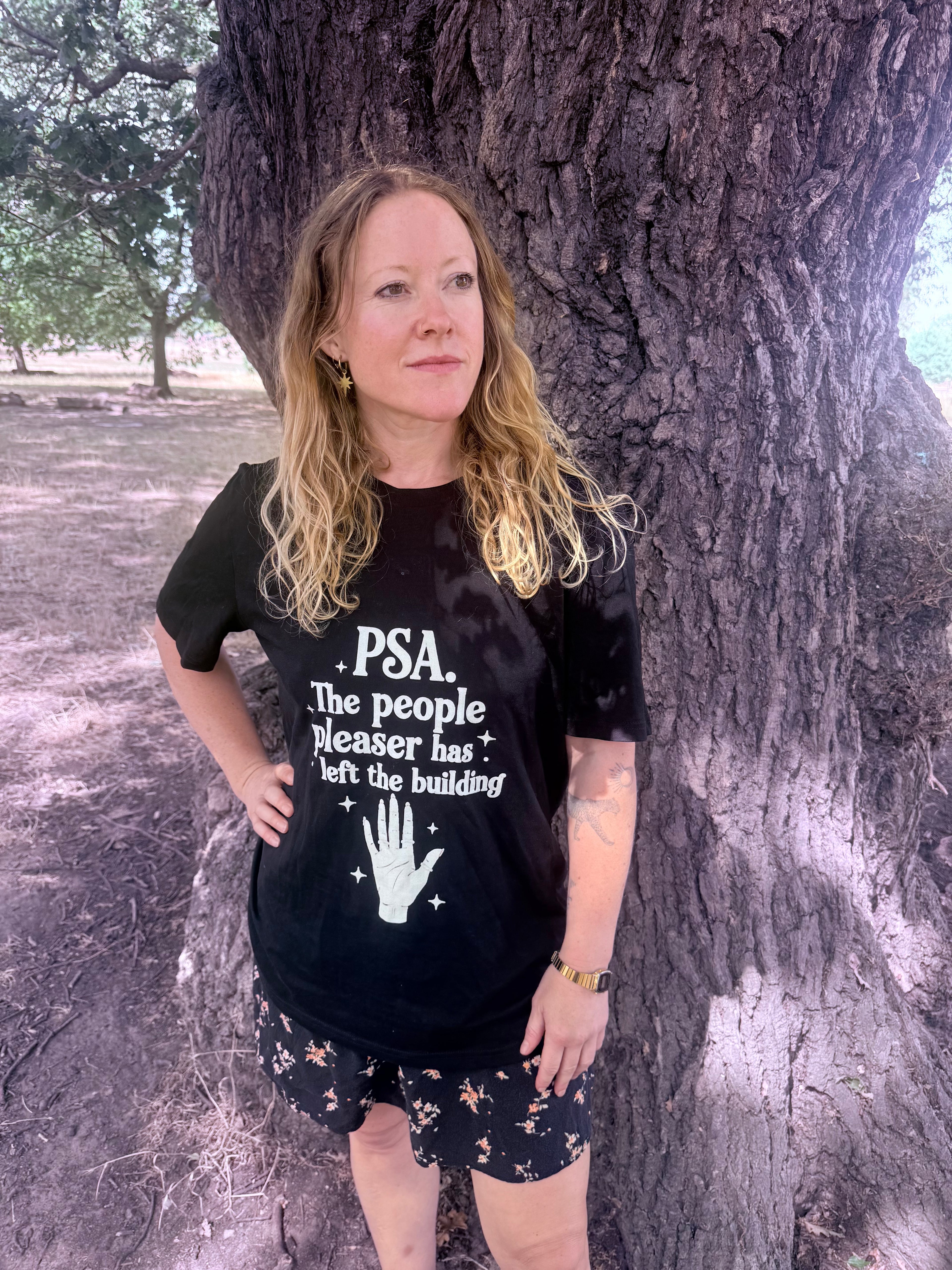 A woman with blonde hair stands outdoors in front of a large tree trunk, wearing a black graphic t-shirt with white text that reads 'PSA. The people pleaser has left the building' and a white handprint graphic, paired with black shorts featuring a small floral pattern.