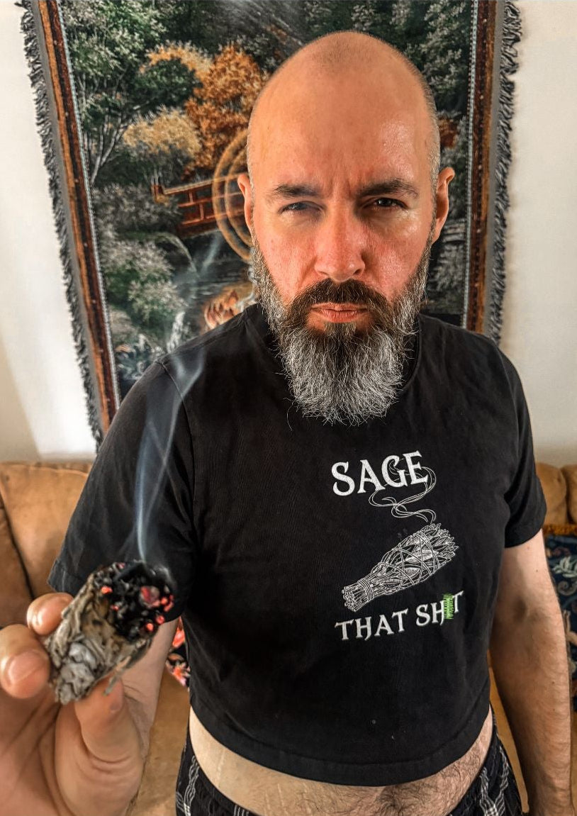 A man with a bald head and gray beard wearing a black graphic t-shirt that reads 'SAGE THAT S...' holds a smoking cone in a home setting with a tapestry in the background.