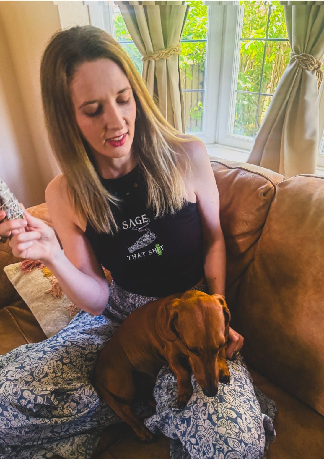 A woman sitting on a brown leather couch with a dachshund dog on her lap, wearing a black tank top with a white graphic print that reads 'SAGE THAT s...'.