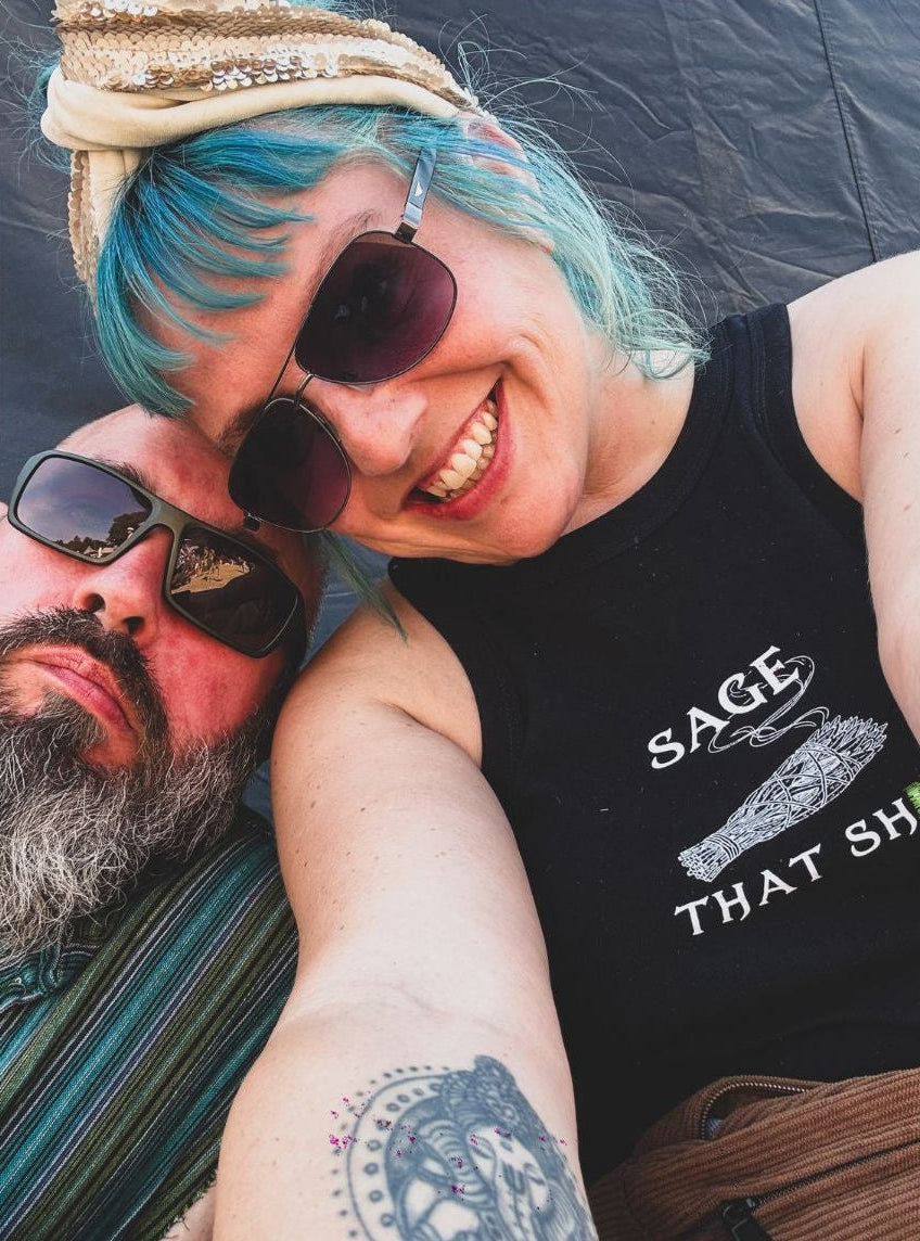 A smiling woman with bright blue hair and sunglasses takes a selfie with a bearded man wearing sunglasses. The woman wears a black tank top with the white text 'SAGE THAT S...'.