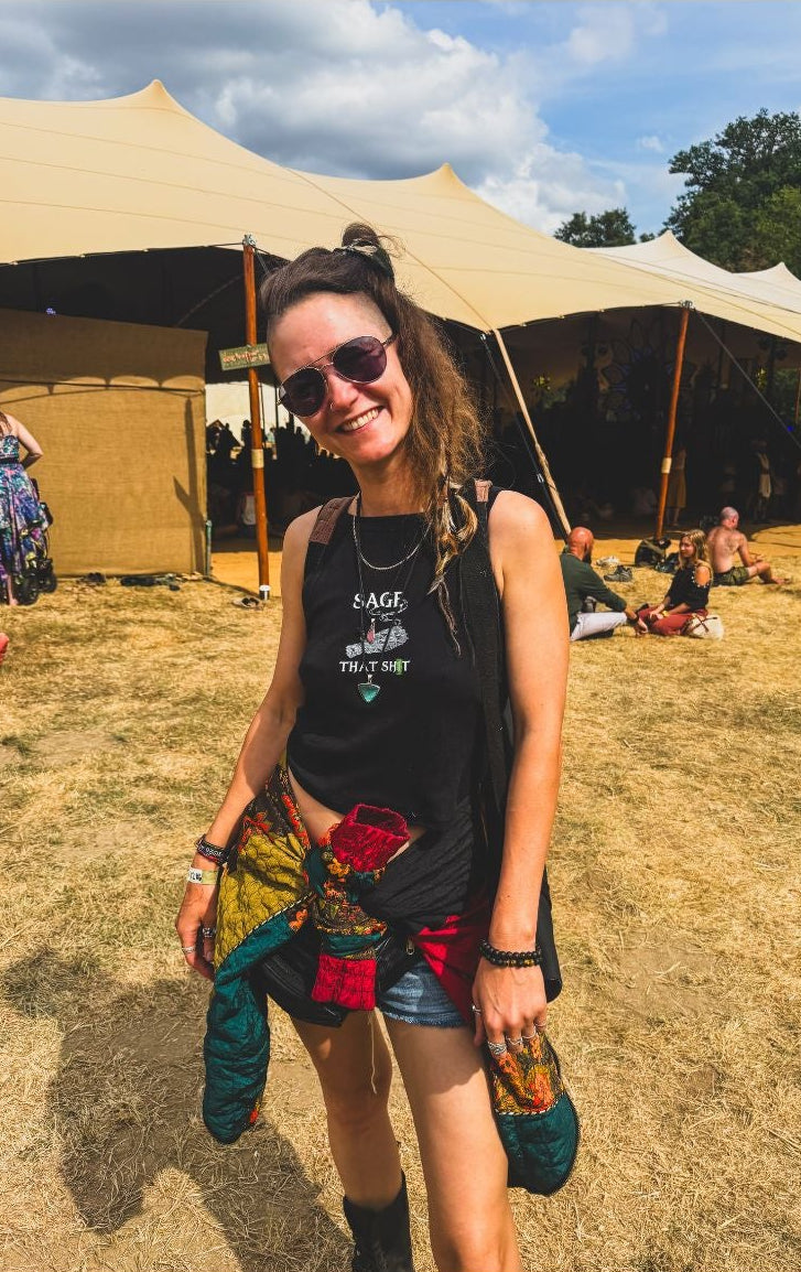 A woman wearing a black graphic tank top that says 'Sage that s...' and sunglasses stands outdoors at a festival or event, holding a colorful patterned garment. Large beige event tents, dry grass, and other people are visible in the background under a blue sky.