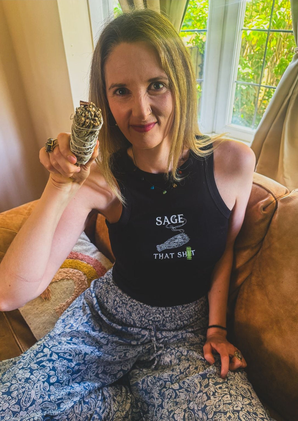 A woman sitting on a brown couch holding a sage smudge bundle, wearing a black tank top with the text 'SAGE THAT S...' and blue floral pajama pants.