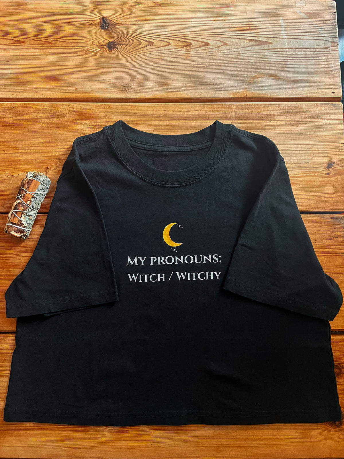 A black short-sleeve t-shirt with a graphic print on a wooden background. The graphic features a yellow crescent moon and the text 'MY PRONOUNS: WITCH / WITCHY' in white.
