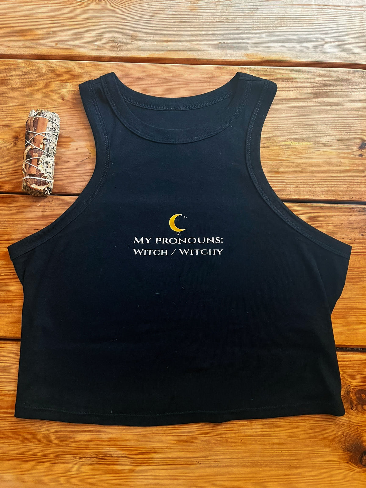 A black sleeveless crop tank top laid flat on a wooden surface, featuring a small yellow crescent moon and white text that reads 'MY PRONOUNS: WITCH / WITCHY' on the chest. A small camouflage-wrapped item sits to the left of the tank top.