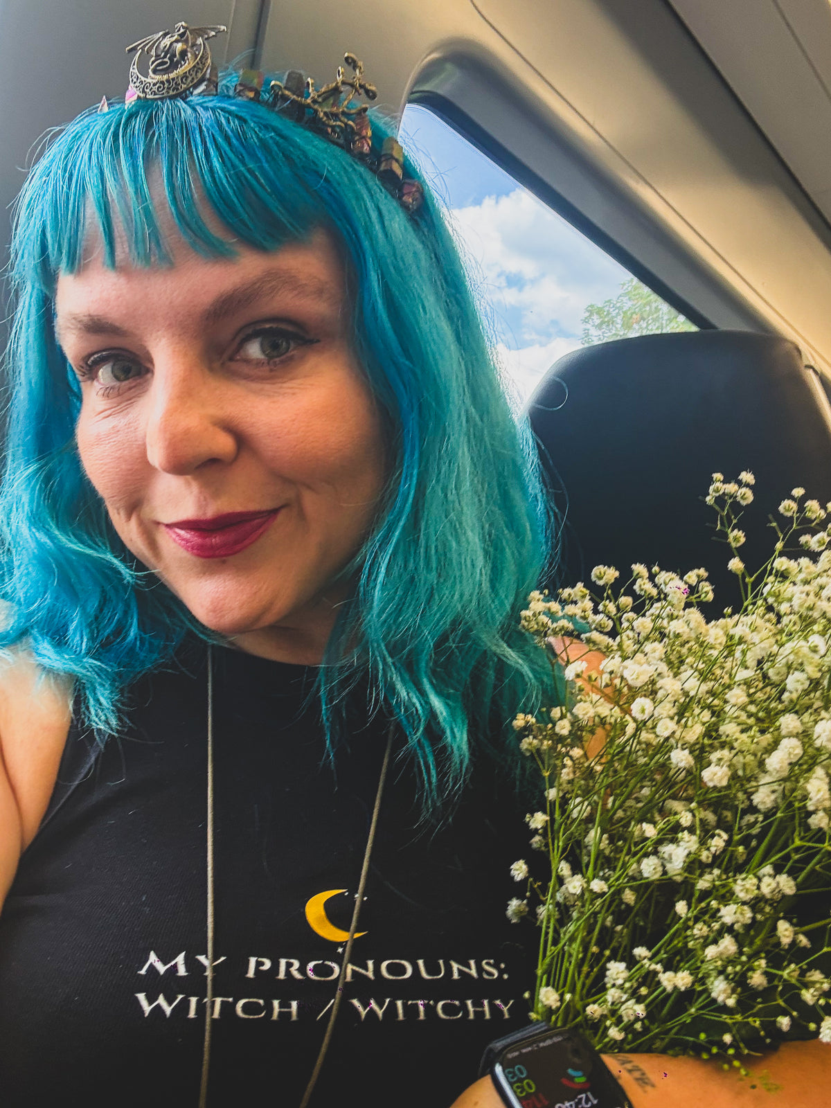 A woman with vibrant turquoise hair wearing a black graphic t-shirt that reads 'MY PRONOUNS: WITCH / WITCHY' with a crescent moon logo. She is sitting in a car, holding a bouquet of white baby's breath flowers.