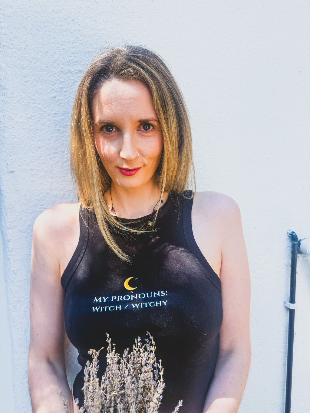 A woman with shoulder-length blonde hair stands against a light blue textured wall, wearing a black tank top with the text 'MY PRONOUNS: WITCH / WITCHY' and a crescent moon graphic on the chest.