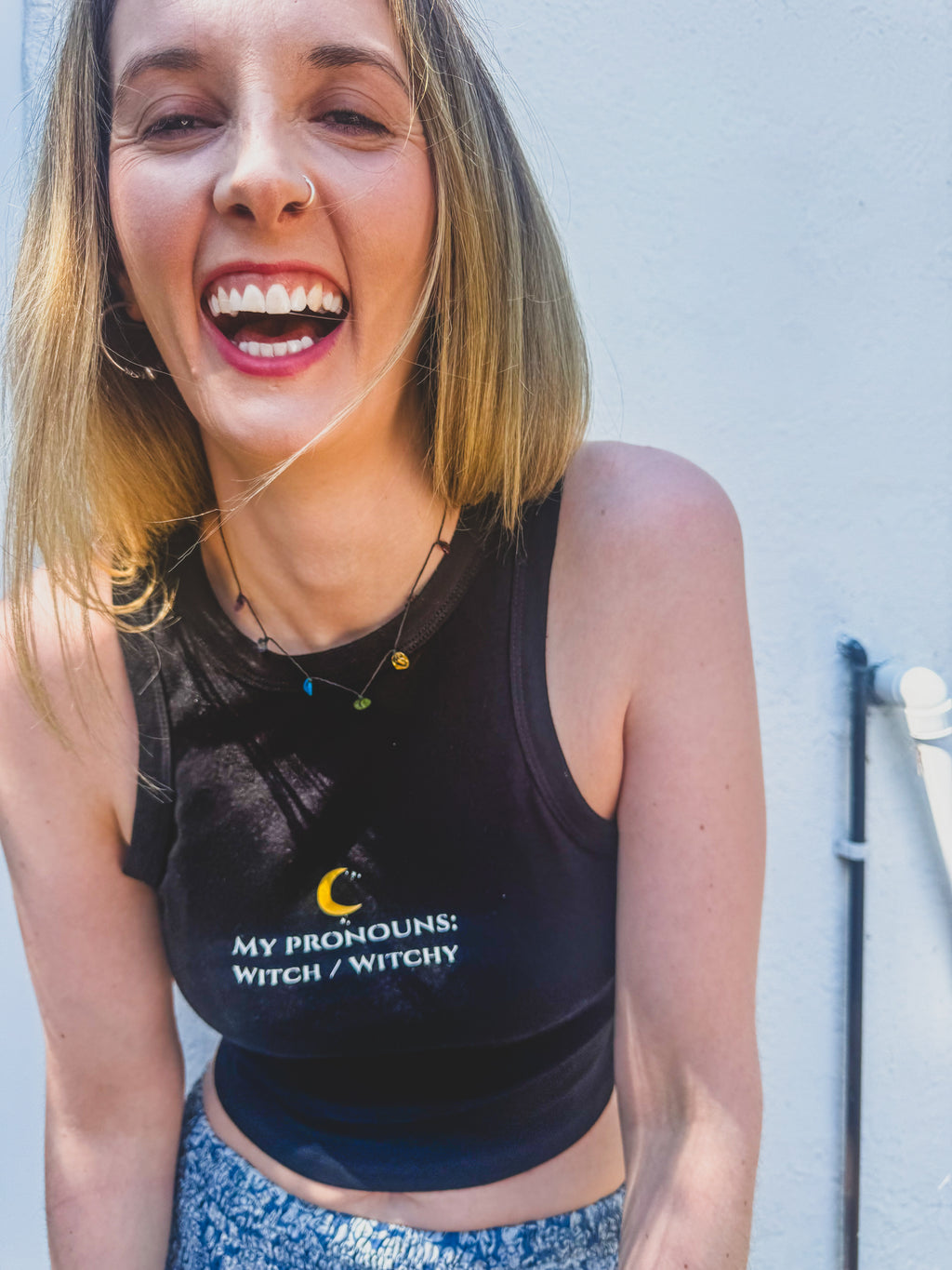 A woman smiling and laughing while wearing a black sleeveless crop top with the text 'MY PROHONS: WITCH / WITCHY' and a small crescent moon graphic on the front, paired with blue patterned bottoms, against a light gray wall.
