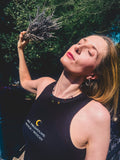 A woman outdoors wearing a black tank top with the text 'MY PRONOUNS: WITCH MYTHICITY' and holding a sprig of dried lavender under bright sunlight.