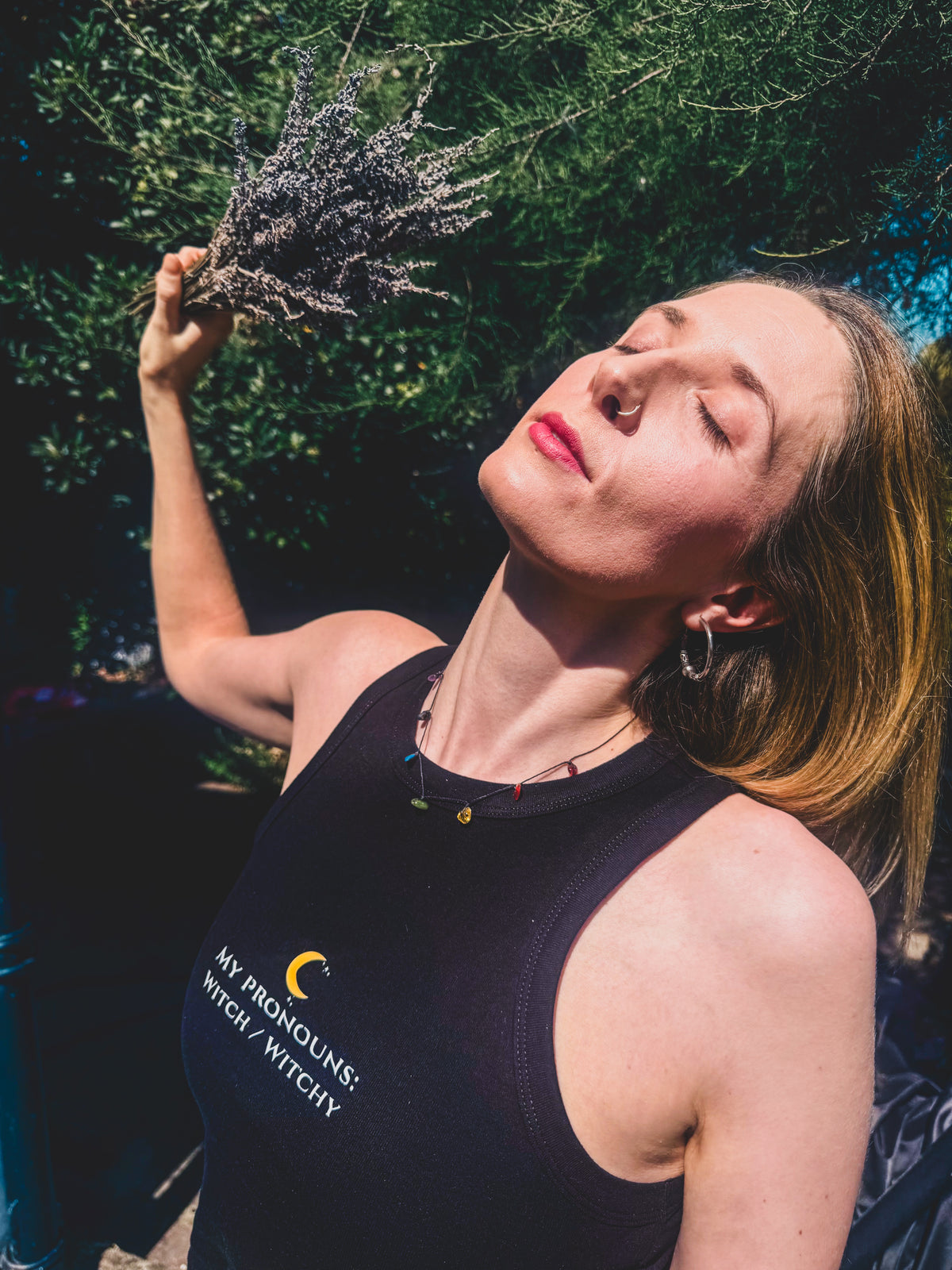 A woman outdoors wearing a black tank top with the text 'MY PRONOUNS: WITCH MYTHICITY' and holding a sprig of dried lavender under bright sunlight.