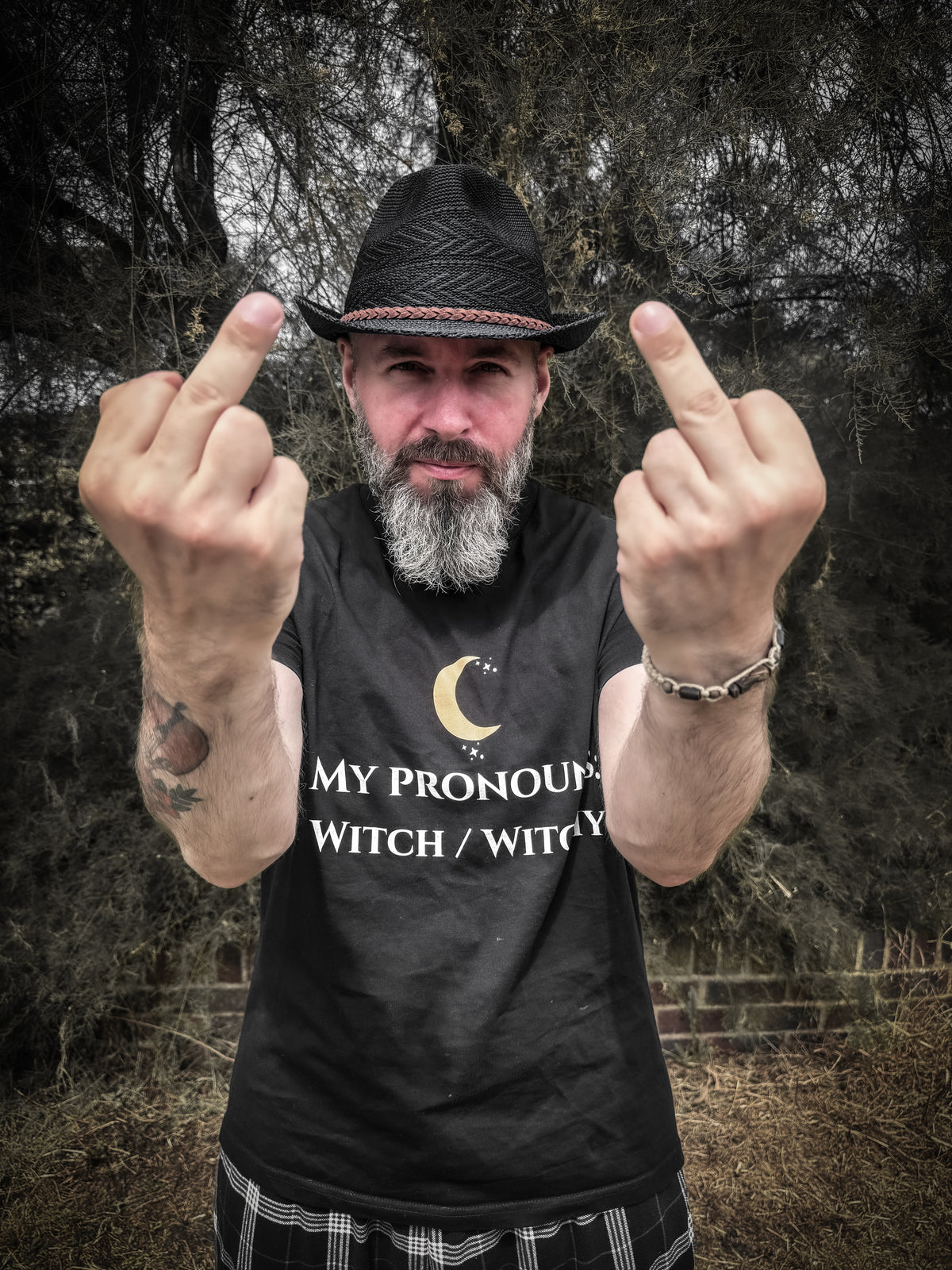 A bearded man wearing a black hat and a black graphic t-shirt with the text 'MY PRONOUND WITCH / WITCH Y' on the front, holding up his middle fingers against a dark, wooded outdoor background.