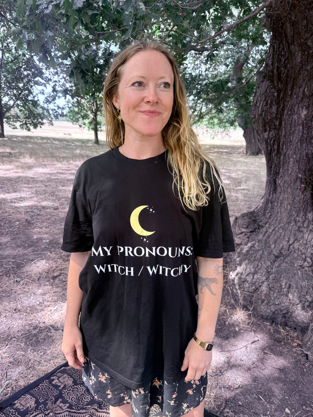 A woman standing outdoors in front of a large tree wearing a black graphic T-shirt with a yellow crescent moon and the text 'MY PRONOUNS WITCH / WITCHY'.