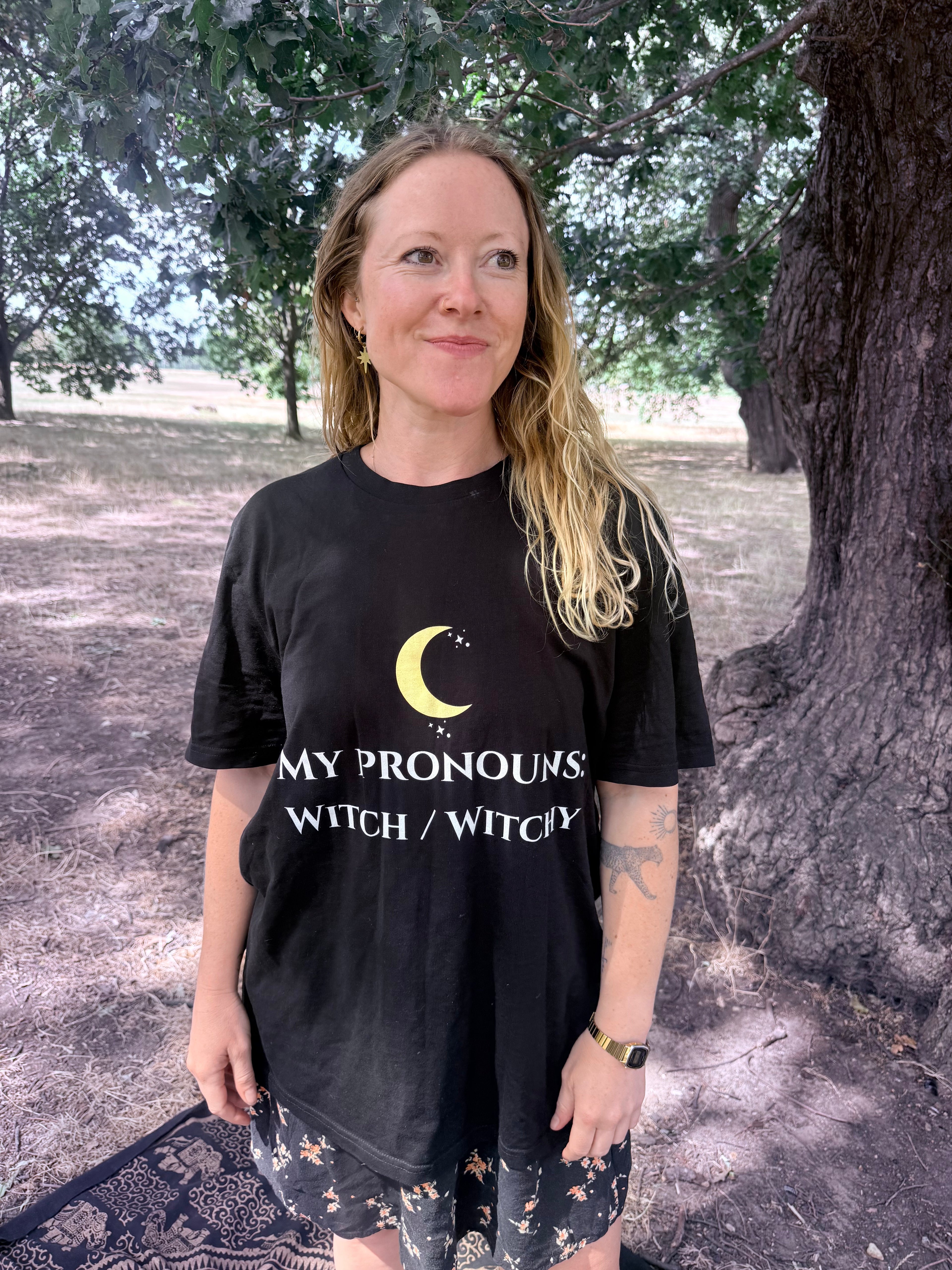 A woman standing outdoors in front of a large tree wearing a black graphic T-shirt with a yellow crescent moon and the text 'MY PRONOUNS WITCH / WITCHY'.